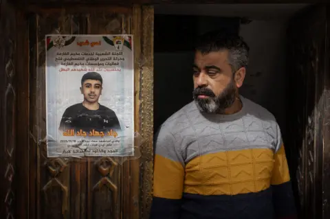 BBC Jad's father stands next to a memorial poster of his son, taped to the family's carved wooden front door in al-Far'a refugee camp, in the occupied West Bank
