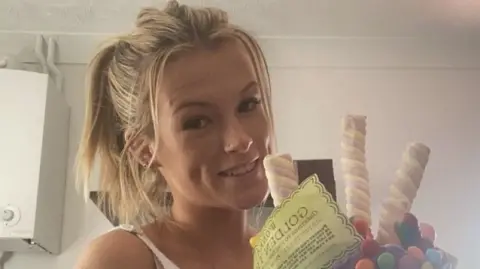 A young blonde woman with her hair tied in a ponytail. She is smiling at the camera and holding a cake covered in Marshmallows, chocolate sweets and a piece of yellow paper which reads "Golden Ticket".