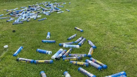 Irene McCormick Blue Cream Deluxe canisters are lying all over a field. They are not in one pile but spread out.