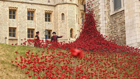 Getty Images Ceramic red poppies positioned on a patch of grass in the Tower of London cascade out from a tall apex of the flowers, with two guards in black uniforms stood nearby.