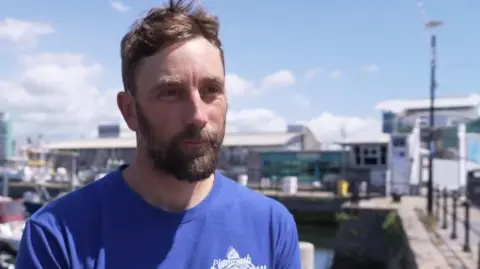A man with brown hair and a beard with a blue t-shirt branded with the Plymouth Fishing & Seafood Association logo. He is standing in front of the harbour