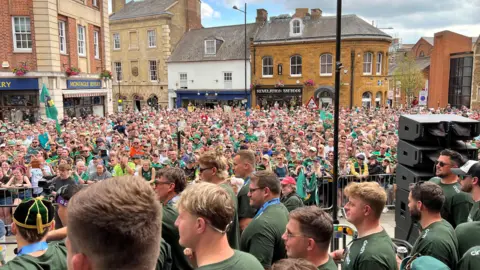 Huge crowds of people lining the streets and cheering on the players - who are wearing green shirts walking in front of the crowd.