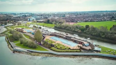 A computer generated image of an aerial view of the refurbished Lido. The site is surrounded by natural water bodies and trees. It features a long rectangular pool with grass around it, and a long building next to it.