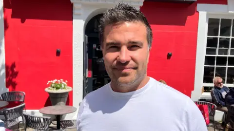 A man is stood in the stood in the sunshine looking at the camera. He has short black hair and a stubbly beard. He is wearing a white t-shirt and it is a head and shoulders shot of him. There are red doors behind him and tables.