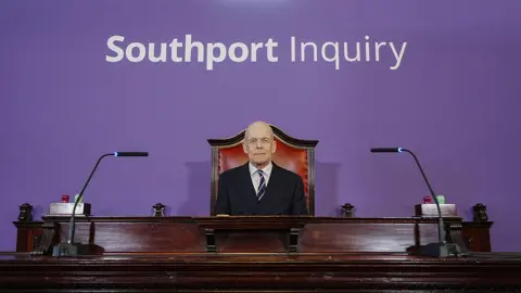 PA Media Chair Sir Adrian Fulford sitting inside the hearing room at Liverpool Town Hall, Liverpool, ahead of the start of the southport inquiry. There is a purple background with Southport Inquiry written in white above his desk with two microphones either side of him on an old-fashioned red and wood chair.