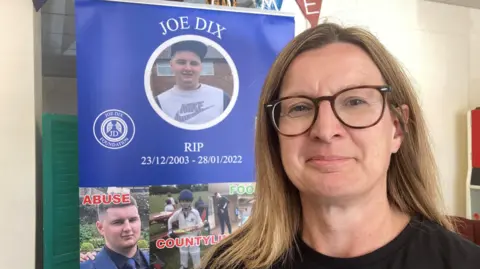 Emma Dix is on the right, looking into the camera and smiling. She is a brunette with shoulder-length hair, and she is wearing rounded glasses and a black t-shirt. To her left, there is a poster for the Joe Dix Foundation, which features images of Joe.
