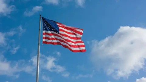 A US flag flying. It has a blue sky behind it and is peppered with clouds