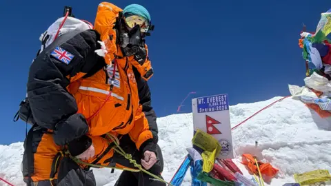 Michael Woods is standing on the side of a snow-covered mountain. He is wearing a High-altitude breathing, a coat with hood up, sunglasses and a hat. 