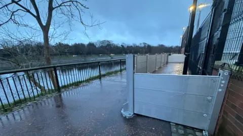 a flood defence barrier along the banks of the Lagan river in Belfast. It is sitting against a building a short distance from riverside railings.