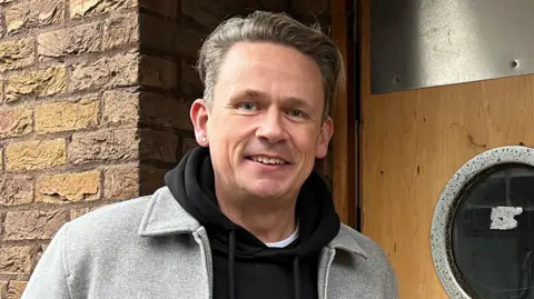 Tom Jackson/BBC Paul Sweeney is standing in front of a door. He has short brown hair and is wearing a black jumper and a grey jacket.