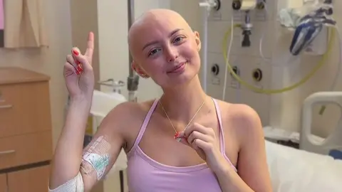 Person sitting in a hospital room wearing a pink top, with a bandaged arm and medical dressing visible. The person holds a necklace while raising one finger. Hospital equipment, tubing, and a bed are visible in the background.