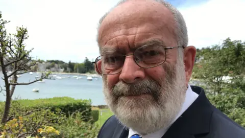 Richard d'Apice, standing outside, by water, with boats on it, with short grey hair and a beard, he is also wearing glasses, a white shirt, with a tie and a blue jacket. His mouth is closed. Foliage is all around him. 