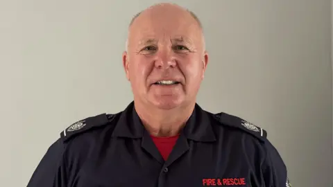 SFRS A man with cropped greying hair smiles at the camera against a light backdrop. He is in navy blue fire service uniform shirt with a red T-shirt underneath.
