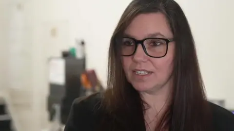 A woman with long dark-brown hair looks towards the camera. She is wearing black-rimmed, large glasses and a smart black jacket.