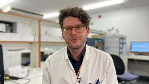 Dr Alex Lawson in his lab coat with an office in the toxicology lab in the background