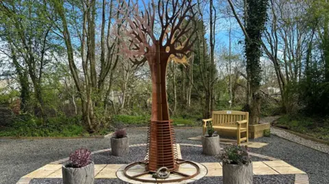 BBC/Charlotte Benton A steel tree in a memorial garden with a bench behind it and a gravel path around it. 