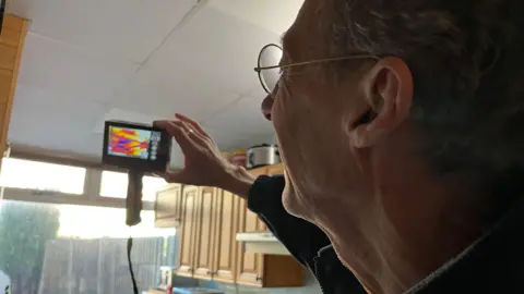 A man holding an infrared camera up near the ceiling in a kitchen, in front of a window. It shows red, yellow and blue shades. He is wearing round glasses and facing away from the camera.