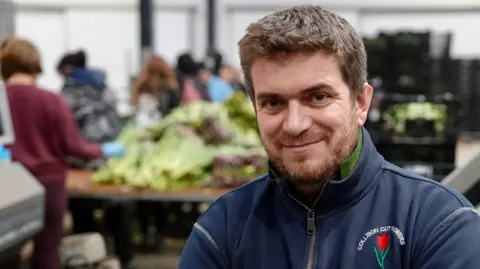 A man with thick brown hair and a short beard smiles with closed lips as he stands in a warehouse where flowers are being processed. He is wearing a blue fleece with a tulip logo and words Collison Cut Flowers. The background is out of focus, but half a dozen people can be seen working at tables covered in flowers. Stacked black crates can also be seen.