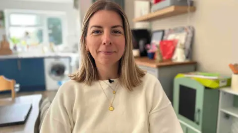 Kate Bradbrook/BBC Anna has fair hair and is wearing a cream jumper with two gold necklaces. She is sitting in a kitchen.