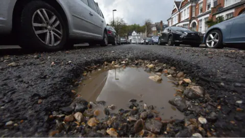 A close-up of a pothole in the middle of a road. There are cars queueing on the left and right hand side of it