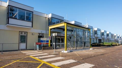 Exterior of the Queen Elizabeth Hospital in King's Lynn - a very long two story building with a walkway and pedestrian crossing on the road in front of it