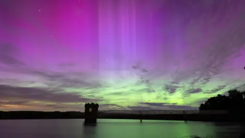 BBC Weather Watchers / lee Northern lights in shades of pink, purple and green are seen above a bridge over water.