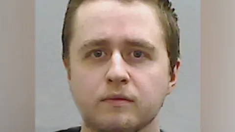 PSNI Alexander McCartney's mug shot. He has short brown hair with a bit of stubble on his cheeks and chin and looking directly at the camera.