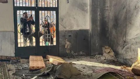 The inside of the mosque, with debris scattered everywhere and black up the walls. Two men are taking photos from the outside through a door.