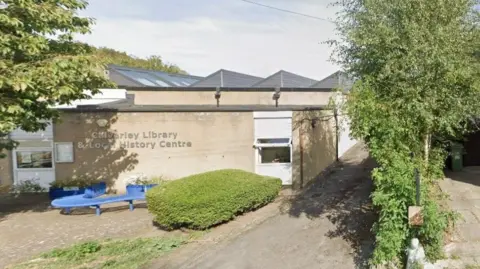 A single-storey, cream-coloured stone building, with a paved area to the front where there is a blue 'S'-shaped bench.