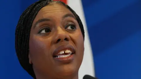 PA Media A close-up shows Kemi Badenoch speaking at a microphone in front of a blue background with part of a Union Jack flag visible behind her. She has black hair in thin braids pinned back from her face.