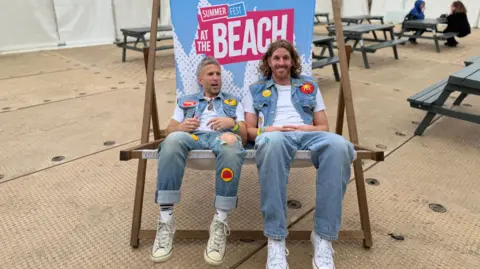 Two men in double denim and white trainers sitting on a giant deck chair at the festival