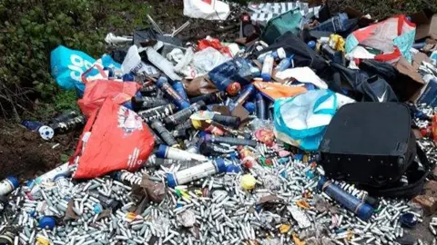 A large selection of rubbish on the ground, with lots of metal canisters. There are bags. Grass is all around it. 