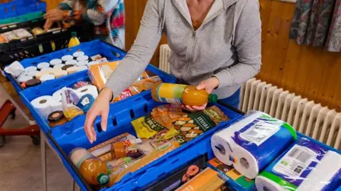 A person in a grey hoodie reaching into a blue basket with groceries inside.