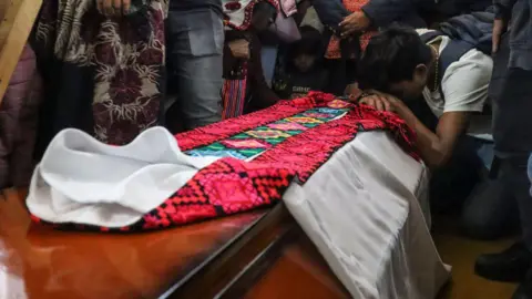 AFP A man is hunched over the coffin of Father Marcelo Perez during his wake. On top of the coffin, the priest's garments are laid out. 