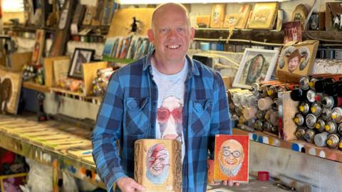 Artist Syd in his workshop holding two paintings of Michael Eavis
