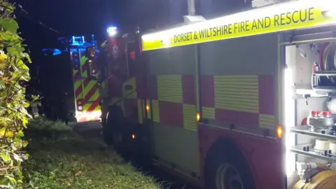 Two Dorset and Wiltshire fire engines parked nose-to-tail in the dark. The hoses and other equipment are illuminated by the on-board lighting.