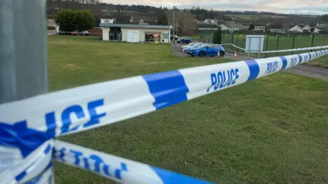 The police tape is close to the camera and can be seen stretching down a road to the car park.