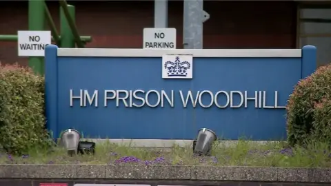 The sign outside HMP Woodhill - a large blue sign sitting between two hedges and on a grassy wall. There is a NO WAITING and NO PARKING sign behind it