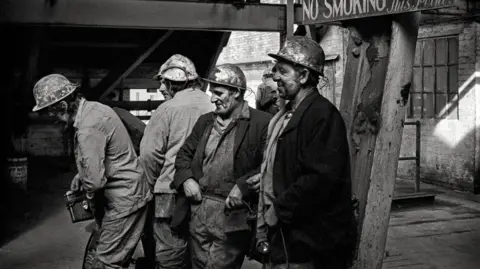 Mik Critchlow A black and white image of a group of miners at the end of their shift. They are all wearing overalls and hard hats and are covered in coal dust.