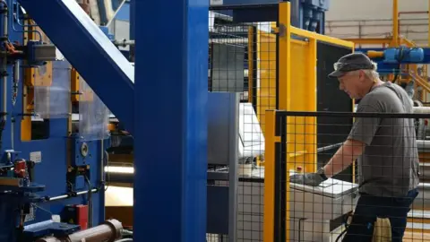 Bridgnorth Aluminium Aluminium worker, wearing cap and safety goggles, standing at a computer with large fabrication machinery around him