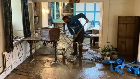 A man in a blue top, wearing boots and holding a broom clears debris from the floor of a flooded pub after the water has receded.