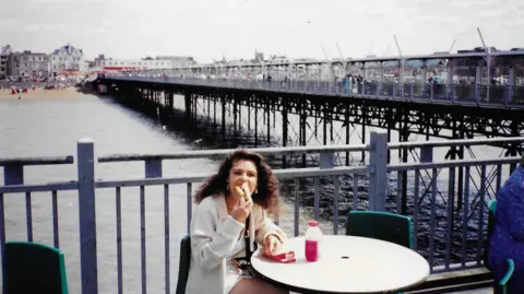 Norman Taylor A photo of a woman with brown curly hair sitting at a table eating a sandwich or burger. She is sitting in front of a pier, with the sea beneath it.
