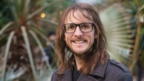 Sheffield Green Party A man with shoulder-length brown hair and a beard. He is wearing glasses and a grey overcoat and stands in front of greenery, smiling