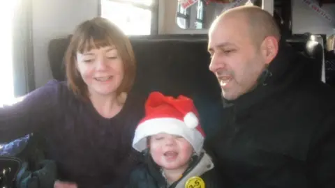 Nicky Wade Nicky Wake is sitting on a train with her husband Andy and her son Finn. It is a candid photograph and Finn is wearing a Santa hat.