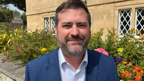 Kevin Guy. He has short dark hair and a beard. He is wearing a checked shirt and a navy blue jacket. He is looking at the camera and smiling.