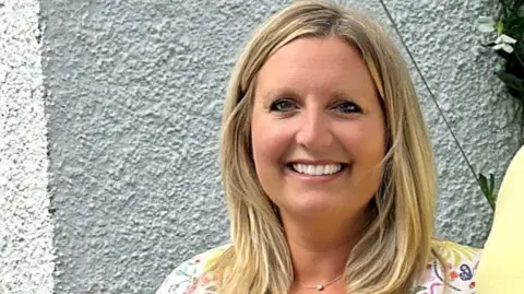 Head and shoulders shot of Nicola Purdie standing against a white wall. She has shoulder length blonde hair and wearing a flowery light coloured top. She is smiling directly at the camera.