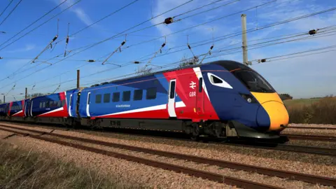 Department for Transport A train with the new red, white and blue branding - it is on a track somewhere in the countryside, with cabling overhead, a blue sky behind it, and fields stretching into the distance. 