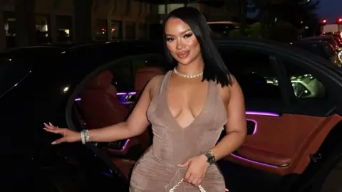 RIELLEUK / Instagram A woman in a light brown dress stands in front of a car, with a shell necklace and straight black hair.