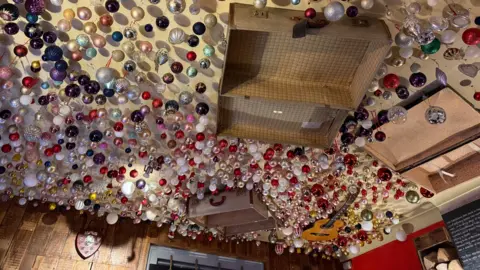 Danny Fullbrook/BBC The ceiling of a pub decorated with hundreds of baubles. It also is decorated with old open suitcases and a guitar.