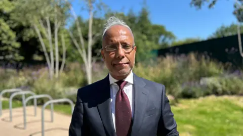 Man wearing a navy suit jacket, white shirt and burgundy patterned tie, has spectacles, and short white hair. Behind him is a bike rack and some lawn and floral borders showing purple flowers blurred out.